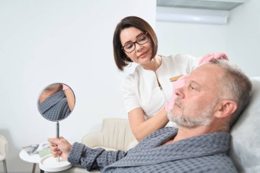 Cosmetologist works with the forehead of an elderly patient, these are beauty injections, man watches the process in the mirror