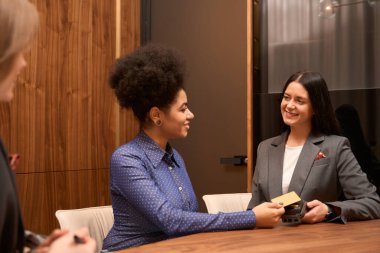 Smiling female sitting by the table and holding credit card, administrator sitting near and holding POS terminal