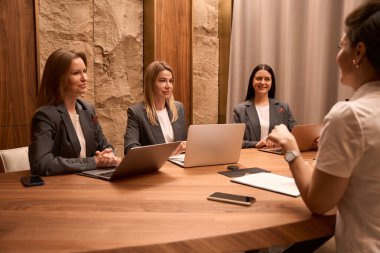 Female doctor in white protective suit, sitting at the table and doing conference with workers in clinic