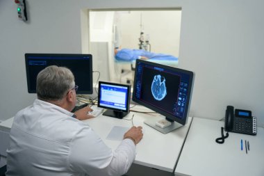 Elderly doctor performs MRI diagnostics in a modern clinic, the patient lies in the tunnel of the apparatus