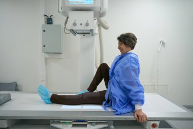 Optimistic elderly woman in special clothes is in the x-ray room, modern equipment is around