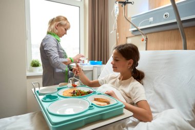 Nurse in medical clothes standing in ward, lady sitting on bed and eating breakfast