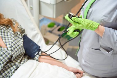 Beautiful woman lying on bed in ward, nurse standing near and measuring blood pressure
