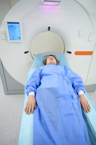Patient, stretching her arms along the body, lies on the mobile surface of the CT machine, she is calm