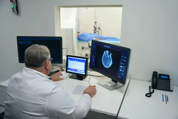 Elderly doctor performs MRI diagnostics in a modern clinic, the patient lies in the tunnel of the apparatus
