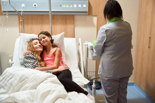 Happy female lying on bed in ward, daughter lying near and hugging her, nurse standing near