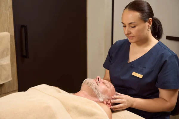 Patient lies on the massage table with his eyes closed, the woman massages his head