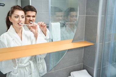 Cute young couple in the bathroom in front of the mirror brushing their teeth, man and woman in bathrobes