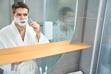 Man in a bathrobe shaves his cheek with razor, he stands in front of wall mirror with an empty shelf