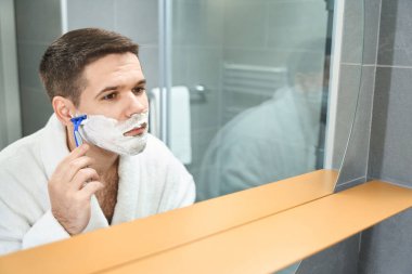 Handsome young man shaves his cheek with a razor, he stands in front of a wall mirror