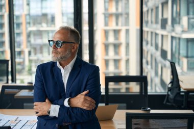 Waist-up photo of determined middle-aged businessman in dark-blue suit standing cross-armed in modern office