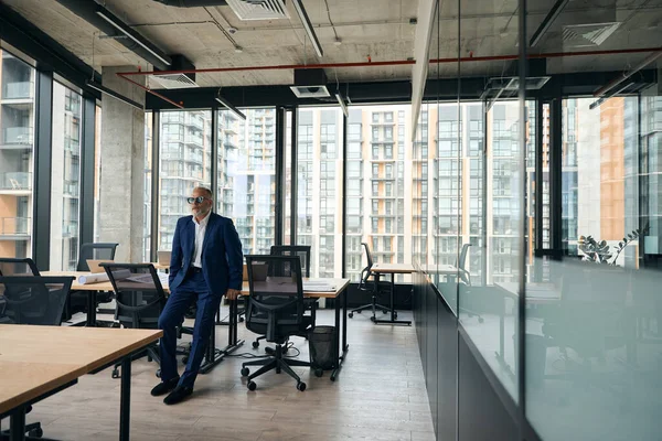 Full-height photo of serious grown entrepreneur leaning on the table in the office while looking to the side