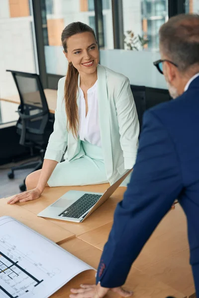 Easy-going businesslady smiling while leaning on the office table and talking to her workspace colleague