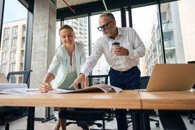 Experienced businessman with cup of coffee in hand pointing to paperwork on table while discussing work project with female colleague