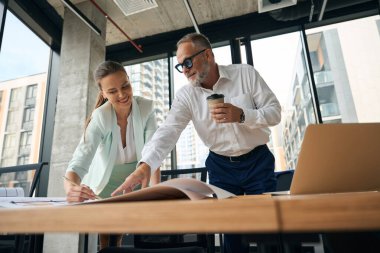 Cheerful businesslady and her thoughtful male colleague looking attentively at spread of paper on the table while working together
