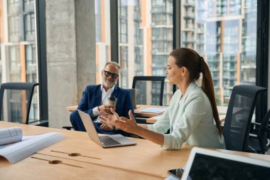 Expressive businesswoman gesturing and sitting in front of laptop at cozy office while middle-aged gentleman listening to her