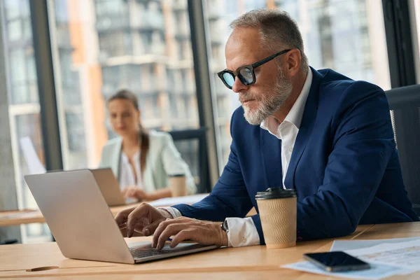 Serious middle-aged office worker looking at computer screen intentionally while sitting at cozy desk with his colleague in the background