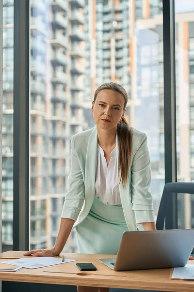 Tired young woman in business-like outfit leaning on her office desk and strictly looking at the camera