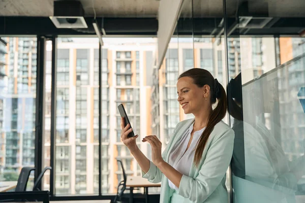Joyous young woman in business suit looking at her smartphone and smiling while standing at well-designed coworking space