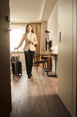 Young woman stands in a hotel room with a travel suitcase, she has a phone in her hands