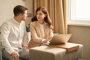 Young woman works on a laptop in a bright room, a man communicates with her