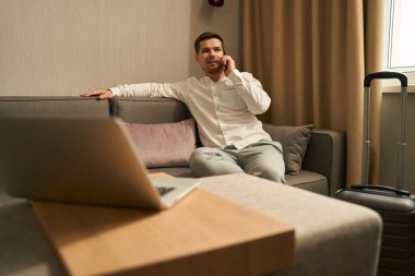 Handsome guy is sitting on the couch and talking on the phone, there is a travel suitcase nearby