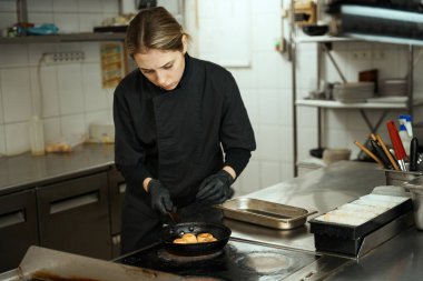 Female chef fries cheesecakes in a cast iron pan on a large stove, a woman in overalls and gloves