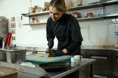 Young woman cuts an orange into circles to prepare a fruit dessert, around the kitchen equipment