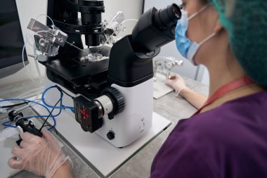Medical worker examines blood samples on a powerful microscope, a woman in a protective mask and gloves