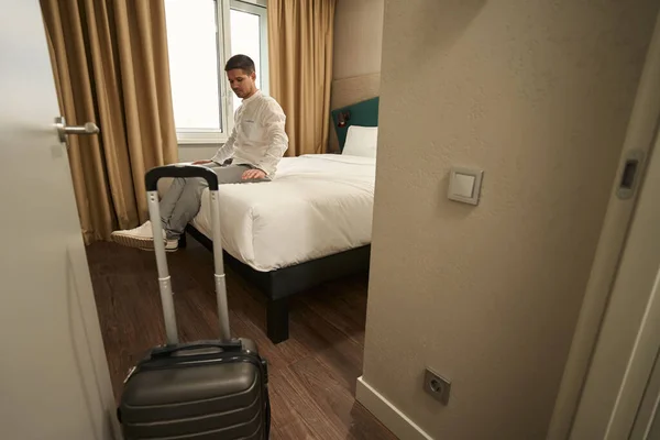 Satisfied Young Man Sitting Soft Bed Hotel Suitcase Standing Nearby — Foto de Stock
