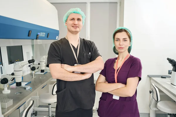 Laboratory staff, a man and a woman, stand indoors at the workplace, arms crossed, they are in overalls
