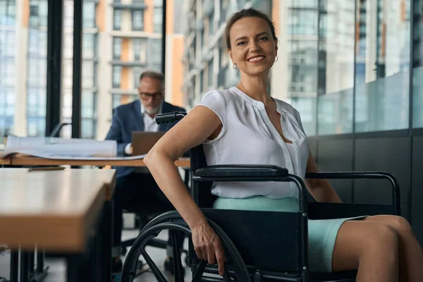 Cheerful woman in a wheelchair is in the office, her colleague is working at a computer