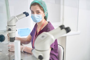 Young laboratory worker is at her workplace at powerful microscope, in background cuvette with liquid nitrogen for vitrification of embryos