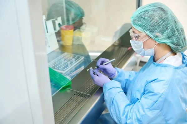 穿着工作服和防护手套的女实验室助理用生物材料在试管上做记号 她在无菌环境中工作 — 图库照片