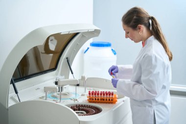 Woman scientist conducts laboratory diagnostics in biochemical analyzer, in front of her are test tubes with blood collected in block