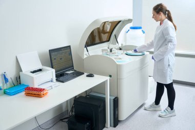 Young employee in overalls stands in front of a modern biochemical analyzer, which is used to obtain kidney samples