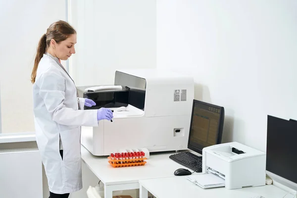 现代化验室的女医生站在血液分析仪旁边 手里拿着一个试管 — 图库照片