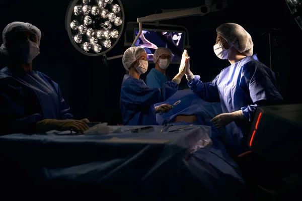 Surgeon and the assistant give high five after a successful operation, the patient lies on the operating table