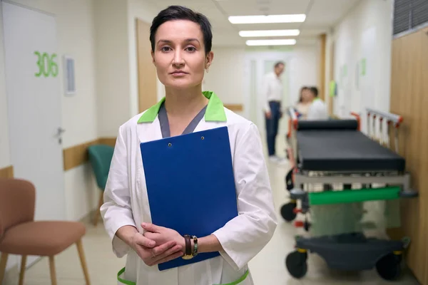 Nurse stands with a blue folder in the corridor of the medical center, in the background people in casual clothes and a mobile bed