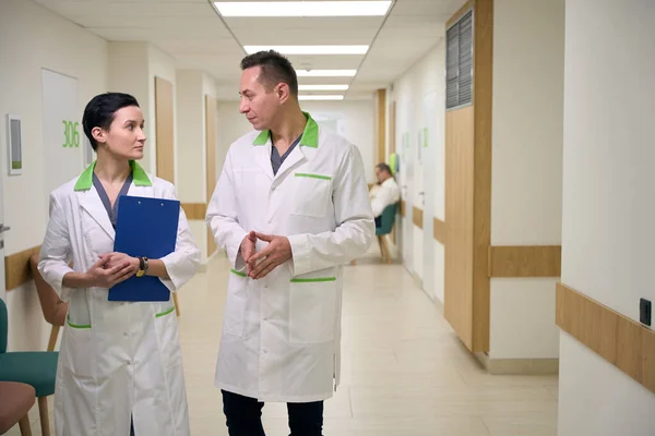 Doctors, man and woman, walk along the corridor ofmedical center, the woman has a blue folder in her hands