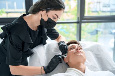 Şırınga tutan ve yakışıklı bir adamın yüzüne güzellik iğnesi yapan koruyucu eldivenli güzel bir doktor.