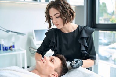 Koruyucu eldivenler giymiş güzel bir kozmetik uzmanı elinde krem tutarak salonda hasta yüzüne ilaç sürüyor.