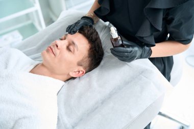 Koruyucu eldivenli hemşire cilt için yağ tutuyor ve salonda insan yüzüne ürün sürüyor.