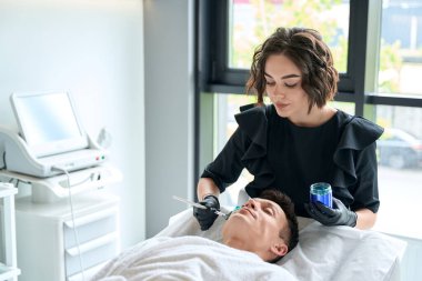 Koruyucu eldivenli güzellik uzmanı elinde deri için maske olan kavanozu tutuyor ve hasta yüzüne fırça sürüyor.