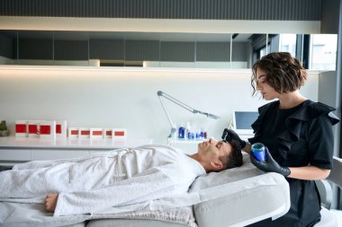 Koruyucu eldivenli kozmetik uzmanı müşterinin yanında oturuyor ve maske takıyor.