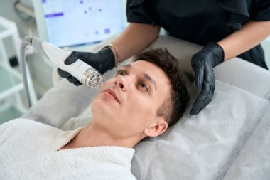 Koruyucu eldivenli bir güzellik uzmanı yetişkin bir erkeğin yanında duruyor ve güzellik salonunda mikro kaldırma yapıyor.