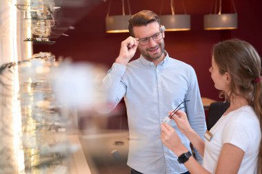 Kendine güvenen yakışıklı erkeğin yakın portresi gözlük çerçevesini denerken gözlükçü dükkanında zarif bir kadınla yeni model hakkında konuşuyor.