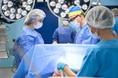 Modern ameliyathane, tıbbi ekip çalışması, insanlar ameliyat önlükleri ve koruyucu maskelerle güçlü lambalar altında çalışıyorlar.