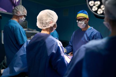 Cerrah, anestezi uzmanı ve asistanlar ameliyat masasında çalışıyor, hasta anestezi altında masada yatıyor.