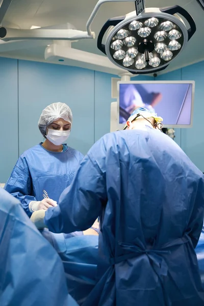 外科医生和助手在一个巨大的特殊灯下操作病人 这个过程在监视器上演示 — 图库照片